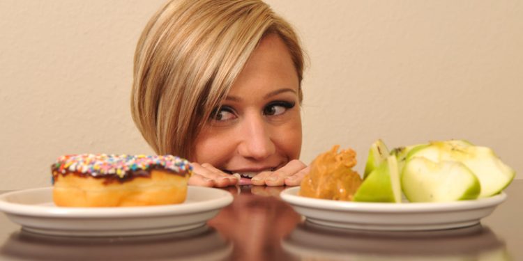 cara de mujer al borde de una mesa mirando un donut con ganas de comerlo y a lado una manzana
