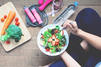 manos sujetan un plato de ensalada y alrededor pesas, metro, agua y cuerda para saltar